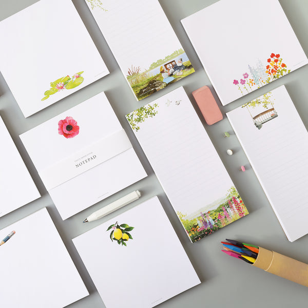 Set of stationery items including our new notepads in their two different sizes, one square and one long for list making. A pen, colored pencils, an eraser, and tacks are between the notepads pads on a gray surface.